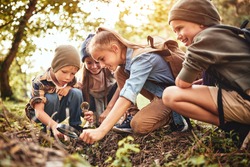 Happy children boys and girls in casual clothes with backpacks making bonfire with magnifying glass together in green forest during school camping activity on sunny day, smiling kids exploring nature