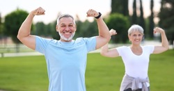 Fit elderly couple working out outside both flexing arms showing biceps symbol of strength looking at camera and smiling, positive husband and wife enjoying morning physical activity on open fresh air
