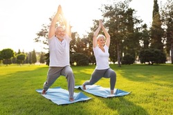 Happy positive fit mature couple of senior woman and man practicing partner yoga on open fresh air outside standing barefoot in warrior Virabhadrasana pose in nature on open air of city park exterior