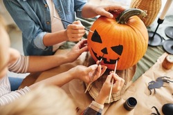 Cropped shot of family parent with little kids preparing for holiday Halloween, hands of mother with children drawing scary face with paint brushes together on pumpkin for house decoration