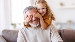 Cute little girl covering eyes with hands of her smiling senior grandfather while playing and having fun together at home, small happy child spending time with positive active grandpa in living room