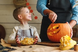 Cute happy little boy helping his father to carve Halloween pumpkin while standing in kitchen at home and preparing for autumn holiday, family of two son and dad making Jack-o-Lantern together