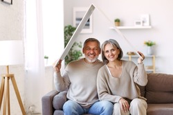 Portrait of happy beautiful senior caucasian family couple holding white roof above head symbol of new home while sitting on sofa, smiling retired husband and wife relocating moving in big dream house