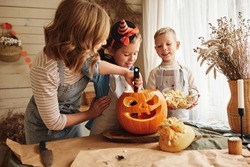 Happy family mother and kids carving pumpkin for Halloween holiday together, preparing for holiday party in kitchen, mom with little daughter and son smiling having fun while creating Jack-o-lantern