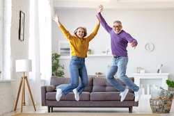 Full length of happy active energetic senior caucasian family couple jumping and having fun together in living room at home, looking at camera and smiling, retired people enjoying life on retirement