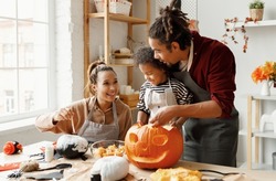 Young joyful african american family mother, father and boy son enjoying Halloween preparation at home, parents and kid making jack o lantern and painting pumpkins for Saints Day celebration