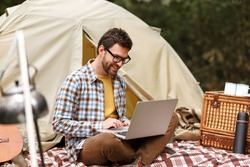 Young smiling man sitting near tent with laptop computer and working remotely while camping in nature, happy male freelancer enjoying remote work at campsite during adventure trip in forest