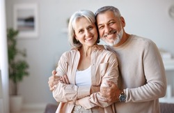 Close up of beautiful smiling senior family couple husband and wife looking at camera with tenderness and love while standing in living room at home, retired man and woman embracing indoors