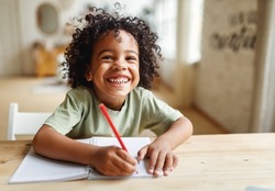 Smiling african american child school boy doing homework while sitting at desk at home, happy mixed race kid practicing handwriting in notebook, learning to write in exercise book