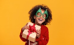 Happy New Year. Portrait of excited little african american boy wearing funny glasses in form of Christmas trees laughing while holding xmas gift box, having fun while standing yellow background