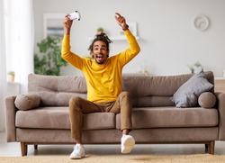 Leisure activities. Young positive happy african american guy with wireless gamepad playing video game while resting at home, sitting in casual clothes on comfortable sofa in cozy living room
