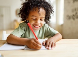 Smiling african american child schoolboy doing homework while sitting at desk at home, happy  kid practicing handwriting in notebook, learning to write in exercise book