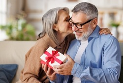 Loving senior woman wife giving  present gift box  for Valentine's day   to happy surprised husband, sitting together on couch at home, smiling mature  man receiving  present. Happy Valentine's day.