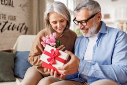 Romantic senior man congratulating happy wife on Valentines Day or March 8 at home, surprised elderly female receiving gift box with red bow and tulips bouquet from loving husband. Selective focus