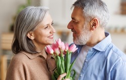 Loving mature husband giving happy wife bouquet of tulips at home, senior man congratulating beloved woman with flowers on March 8. Lovely elderly couple celebrate anniversary. Selective focus