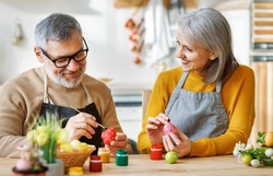 Happy senior couple wife and husband sit at table in kitchen, dye and decorate boiled eggs with food paints, mature family prepare together for Easter holiday and joyfully talking, selective focus
