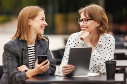 Two young successful businesswomen sitting at outdoor cafe and looking at digital tablet and smartphone with happy face expression during meeting, excited business colleagues feeling sitisfied  