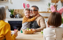 Happy big multi-generation family grandparents, parents and children prepare for Easter holiday at home together, grandfather and grandson in bunny ears hugging while painting eggs in kitchen