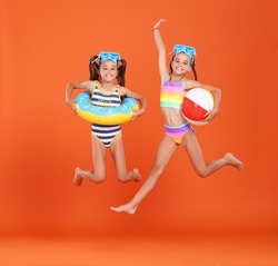 Two funny little girls sisters in swimsuits and swimming goggles jumping up in air with inflatable swimming ring and ball in hands on orange studio background, excited and ready for summer   vacation