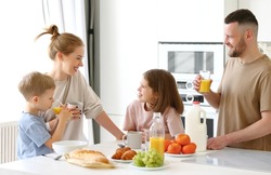 Young happy beautiful family having breakfast together at home. Father, mother and two cute little kids eating healthy food in morning, talking and smiling while standing in modern kitchen