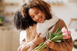 Little african american son congratulating mom with Mother's day at home and giving her fresh flower bouquet, happy mixed race woman mother embracing son. Family holidays and celebration concept