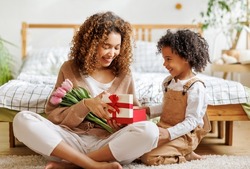 Cheerful family mom opens a gift box with her son and holding bouquet  of flowers while resting on floor by bed during holiday celebration mothers day at home