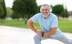 Happy positive mature sportsman during outdoor workout, senior man wearing headphones and sports outfit warming up muscles, doing side squat on one leg, enjoying active lifestyle outside in park