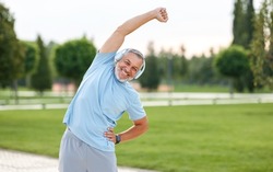Staying active after retirement. Happy joyful mature retired sportsman wearing headphones and sportswear doing side stretching exercises with arm over his head, exercising outside in city park