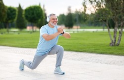 Full-length photo of happy smiling retired sportsman in sportswear and headphones doing front squat on one leg forward with hands folded outside in city park during routine workout in morning