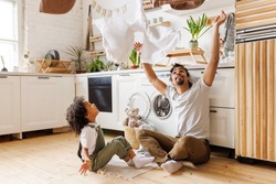 Happy african american dad and son smiling and tossing towels in air while sitting on floor near washing machine during household routine in kitchen at home