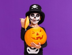 Happy cheerful boy in skeleton costume with  pumpkin  jack-o-lantern  celebrates Halloween and laughs on  bright purple background