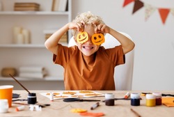 Portrait of cute kid  boy  making Halloween home decorations   while sitting at wooden table, child covers eyes with carved pumpkins and laughs merrily