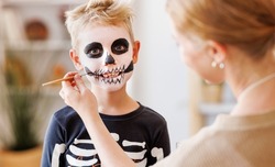 Festive makeup for Halloween. Woman doing skeleton make-up    for a little cheerful boy in costume while preparing holiday at home.