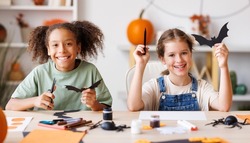 Happy multinational children girls making Halloween home decorations together, kids painting pumpkins and making paper cuttings