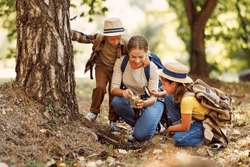 Happy family: two   kids boy and girl  with backpacks looking examining environment through magnifying glass while exploring forest nature  on sunny day during outdoor ecology school lesson