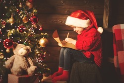 happy child girl writes letter to Santa Claus at the Christmas tree in the evening