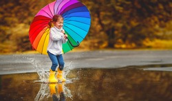 Happy funny ba child by girl with a multicolored umbrella jumping on puddles in rubber boots and laughing