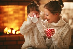 family mother and child daughter drinking tea and laughing on winter evening by fireplace