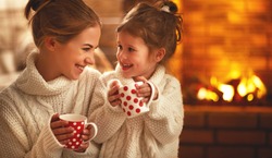 family mother and child daughter drinking tea and laughing on winter evening by fireplace