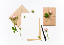 Letter, envelope and a present in eco paper on white background. Wedding invitation cards or love letter with pink roses. Valentine's day or other holiday concept, top view, flat lay, overhead view
