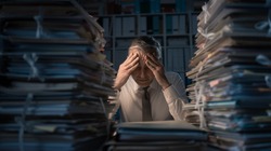 Desperate businessman working in the office late at night and overloaded with work, his desktop is covered with paperwork: business management and deadlines concept
