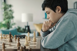 Focused smart boy playing chess at home, he is thinking and looking at the chessboard
