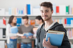 Confident handsome student holding books and smiling at camera, library bookshelves on background, learning and education concept