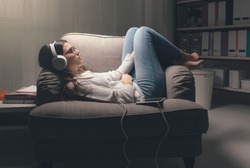 Young woman with headphones relaxing at home late at night, she is lying on the armchair and listening to music using a tablet