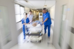 A motion blurred photograph of a patient on stretcher or gurney being pushed at speed through a hospital corridor by doctors & nurses to an emergency room