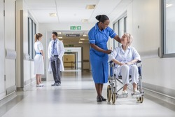 Senior female woman patient in wheelchair sitting in hospital corridor with African American female nurse doctor and nurse in background