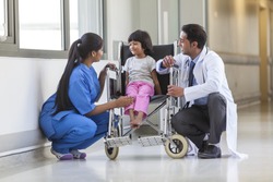 Young female child patient in wheelchair sitting in hospital corridor with Indian Asian female nurse and male doctor