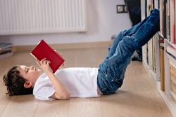Little boy child reading a book in the library. He lies on the floor. Legs on bookshelf