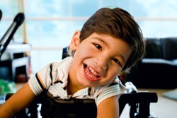 Disabled little boy standing in walker smiling and happily looking at camera with bright blue sky and clouds in the background window