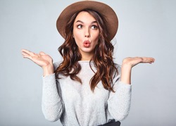 Comparison concept. Young brunette woman in casual clothes and brown hat displaying something on both flat hands for similar choice of product, gray background studio